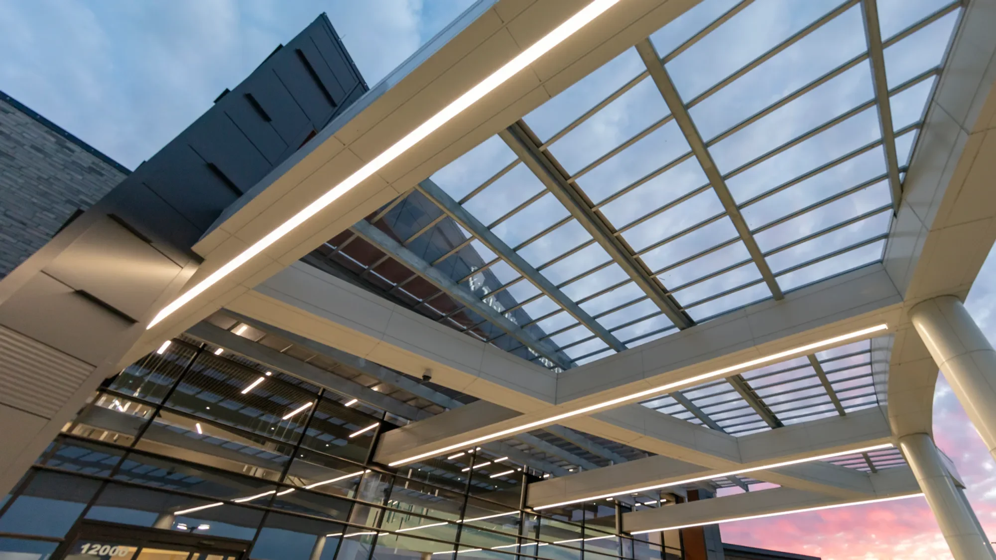 Modern multi-story building with glass windows and a glass roof, illuminated with white lights, taken during twilight with the sky showing shades of pink and blue.