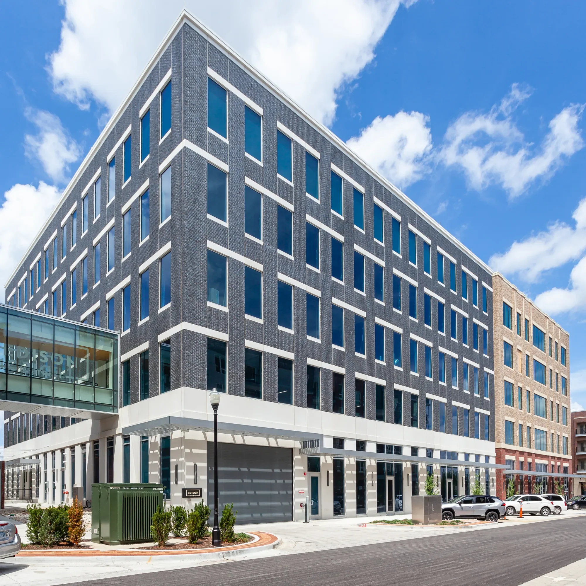 Modern multi-story office building with a dark brick facade and numerous windows, situated in an urban area with cars parked in front and a clear blue sky with some clouds overhead.