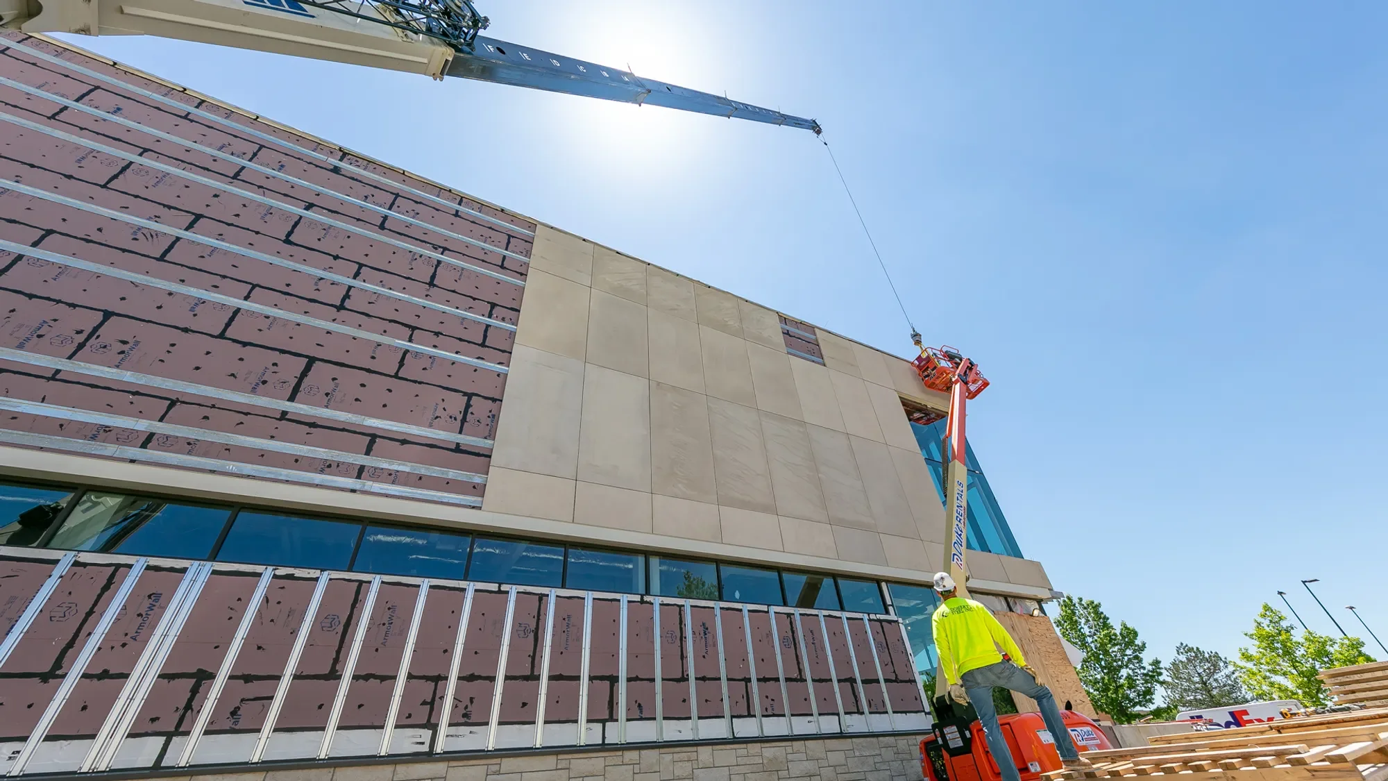 A construction site with workers installing exterior wall panels on a modern building under a clear blue sky. A crane and a lift are in use, and a worker is standing nearby wearing a yellow safety vest.