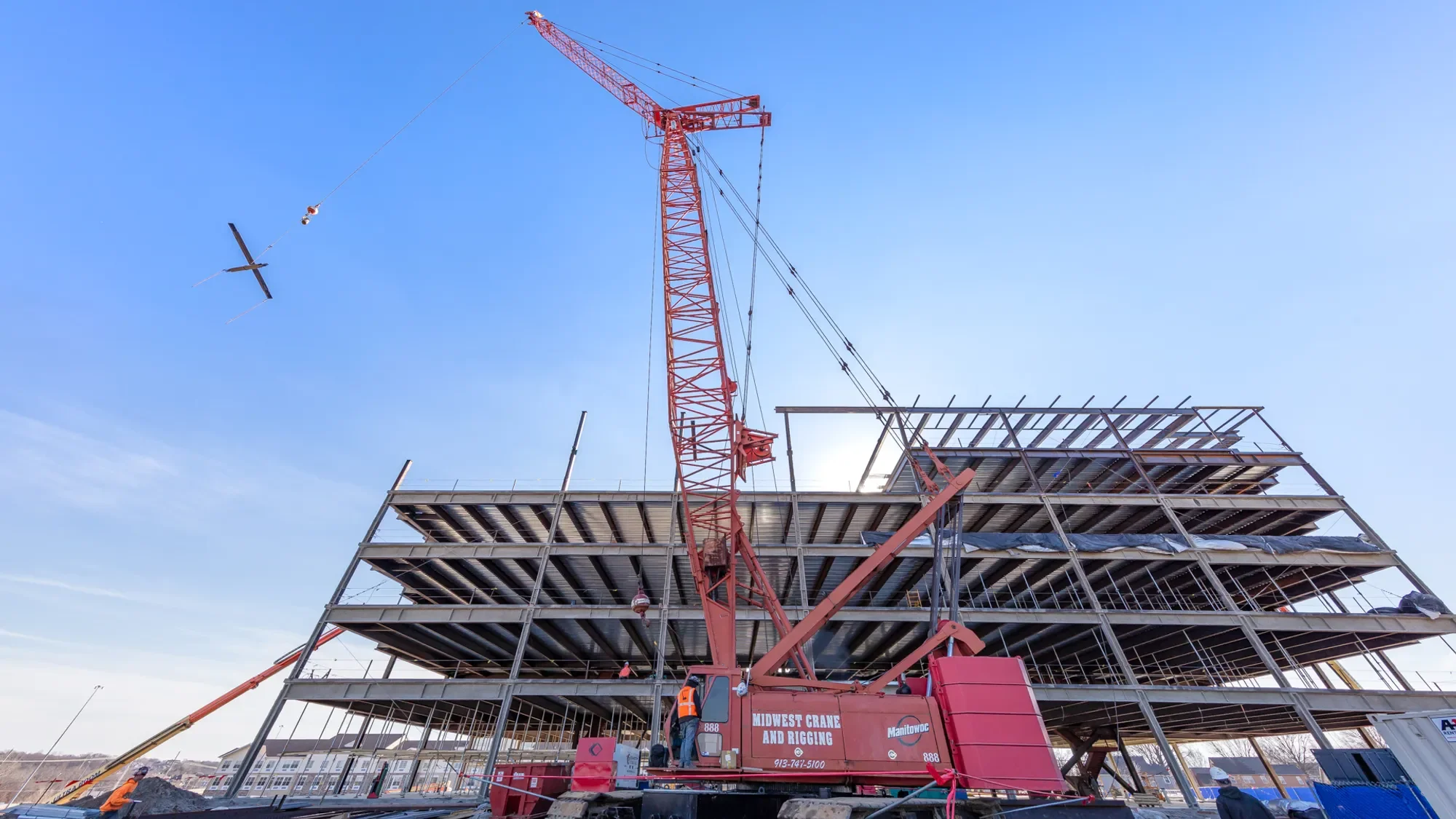 Construction site with a partially built multi-story building and a red crane lifting materials against a blue sky.