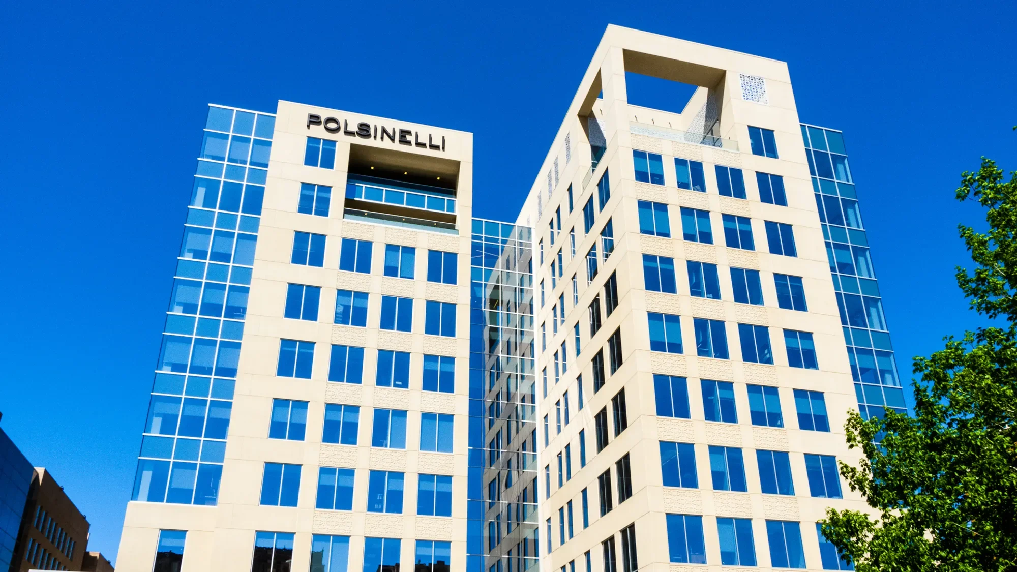 Modern multi-story office building with blue-tinted windows and white exterior, several floors, against a bright blue sky with some trees visible on the right.