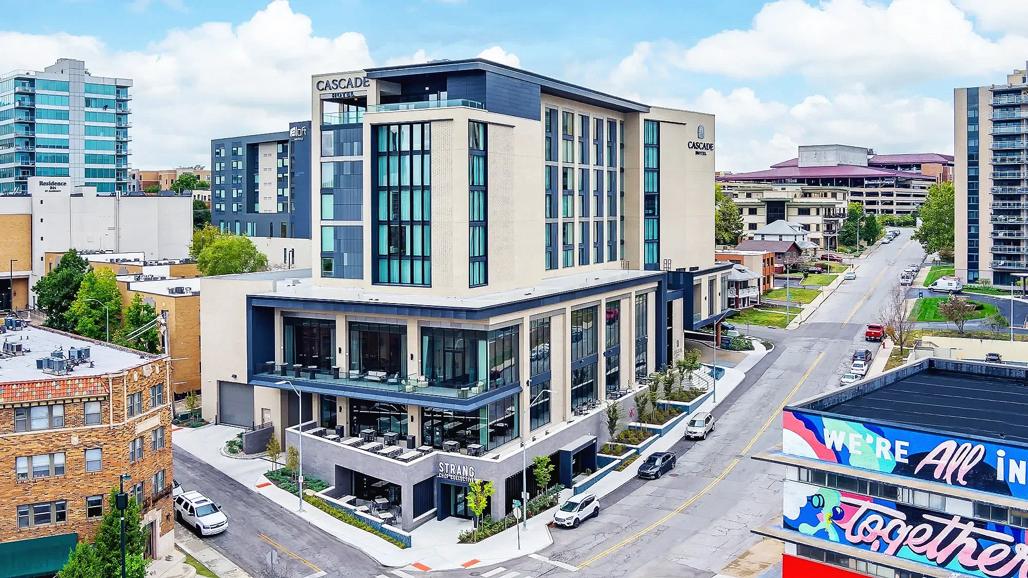 Modern multi-story hotel called with blue and beige exterior, large glass windows, and a rooftop balcony. Surrounding buildings include high-rise and low-rise structures, with a street lined with parked cars, trees, and a billboard with colorful text.