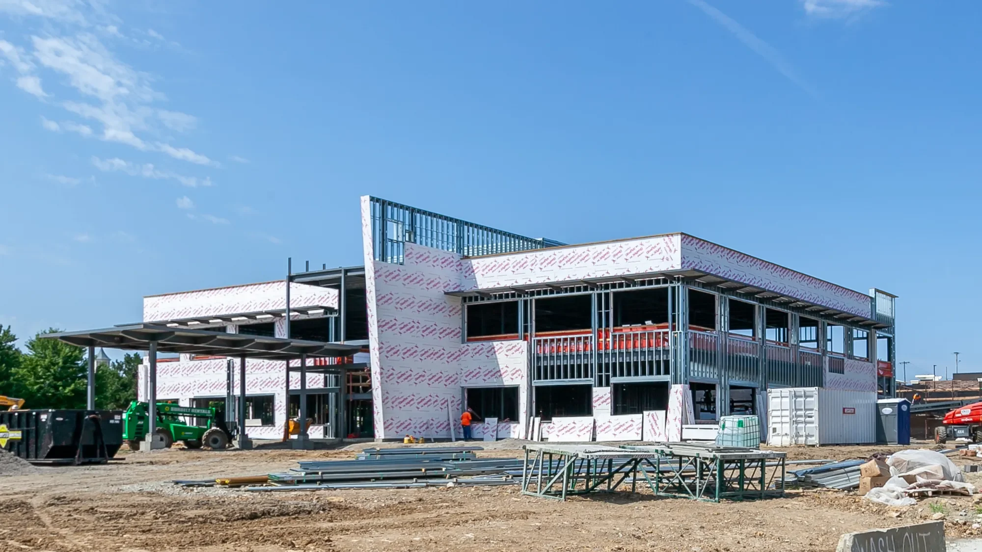 Construction site with a partially built multi-story building covered in building wrap, with scaffolding and construction equipment around, and workers on site under a blue sky.