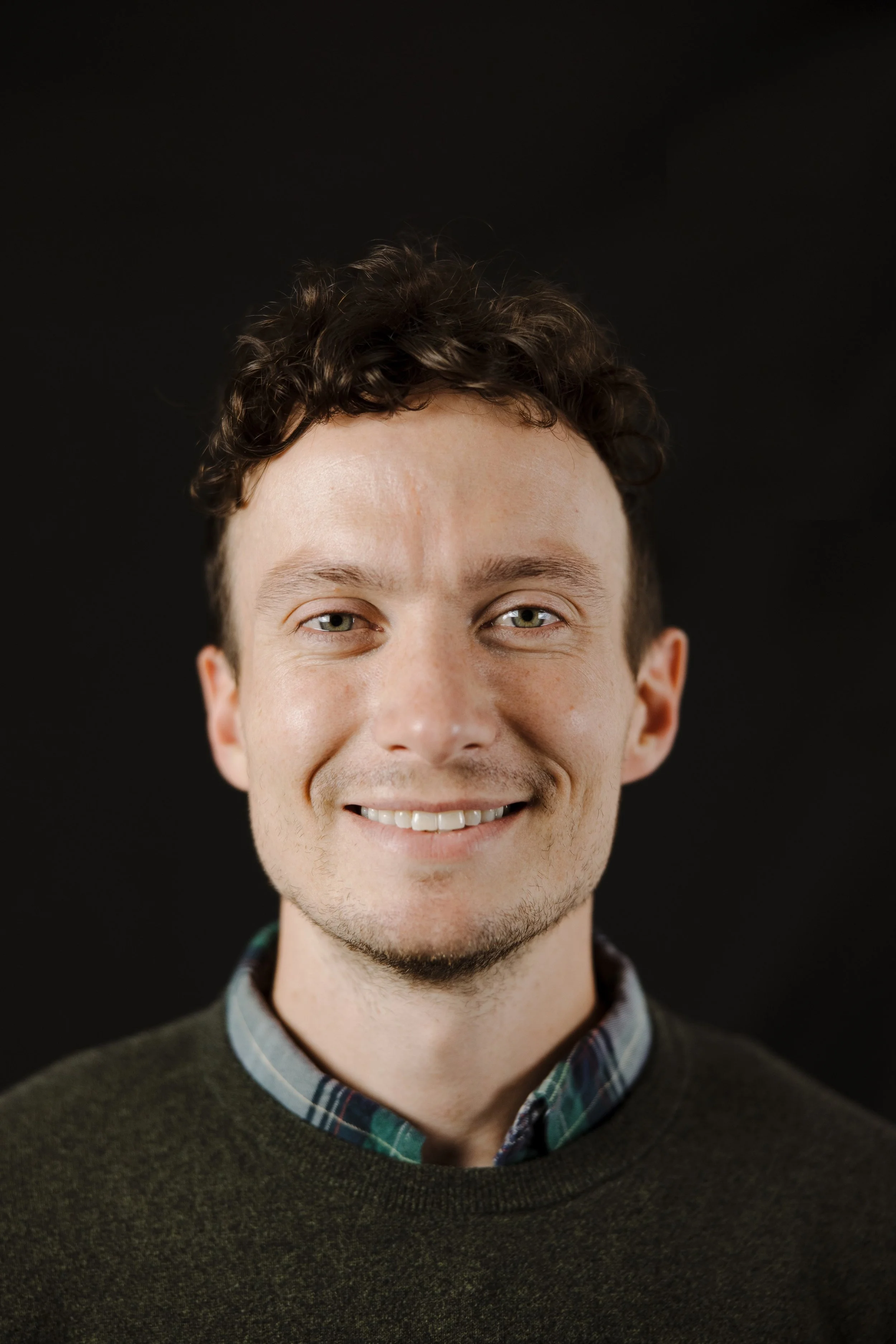 Close-up portrait of a smiling young man with curly brown hair, wearing a dark sweater over a checkered shirt, against a black background.
