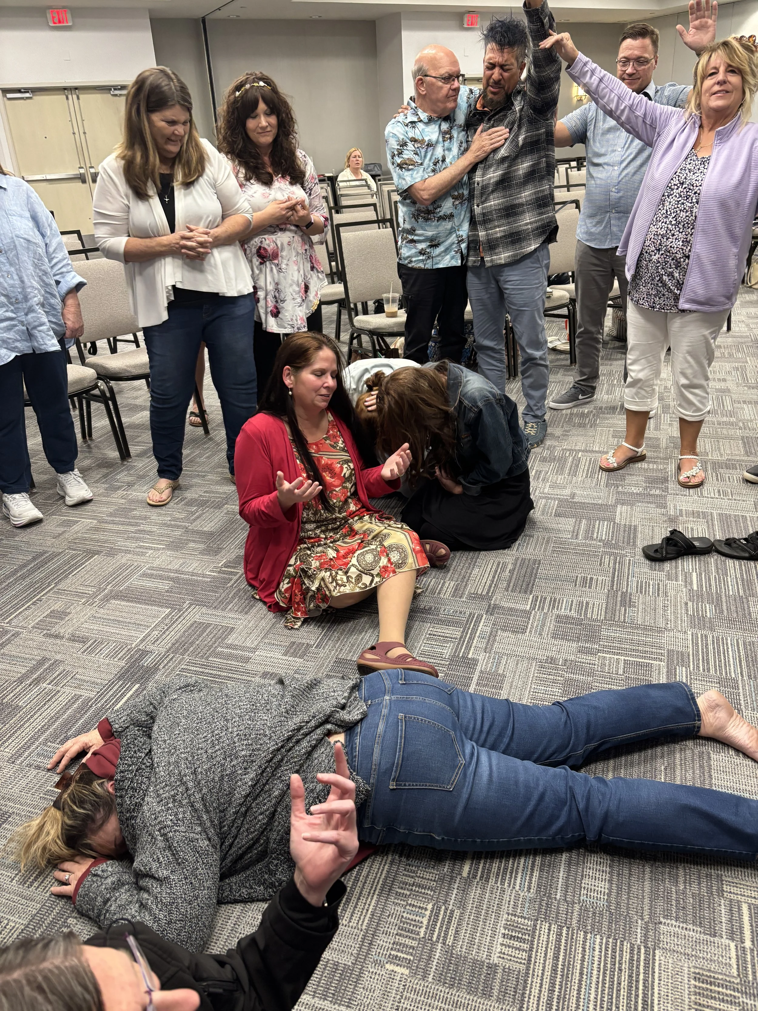 People in a room appear to be praying or experiencing a religious moment, with some kneeling or lying on the floor and others standing with hands raised or clasped.