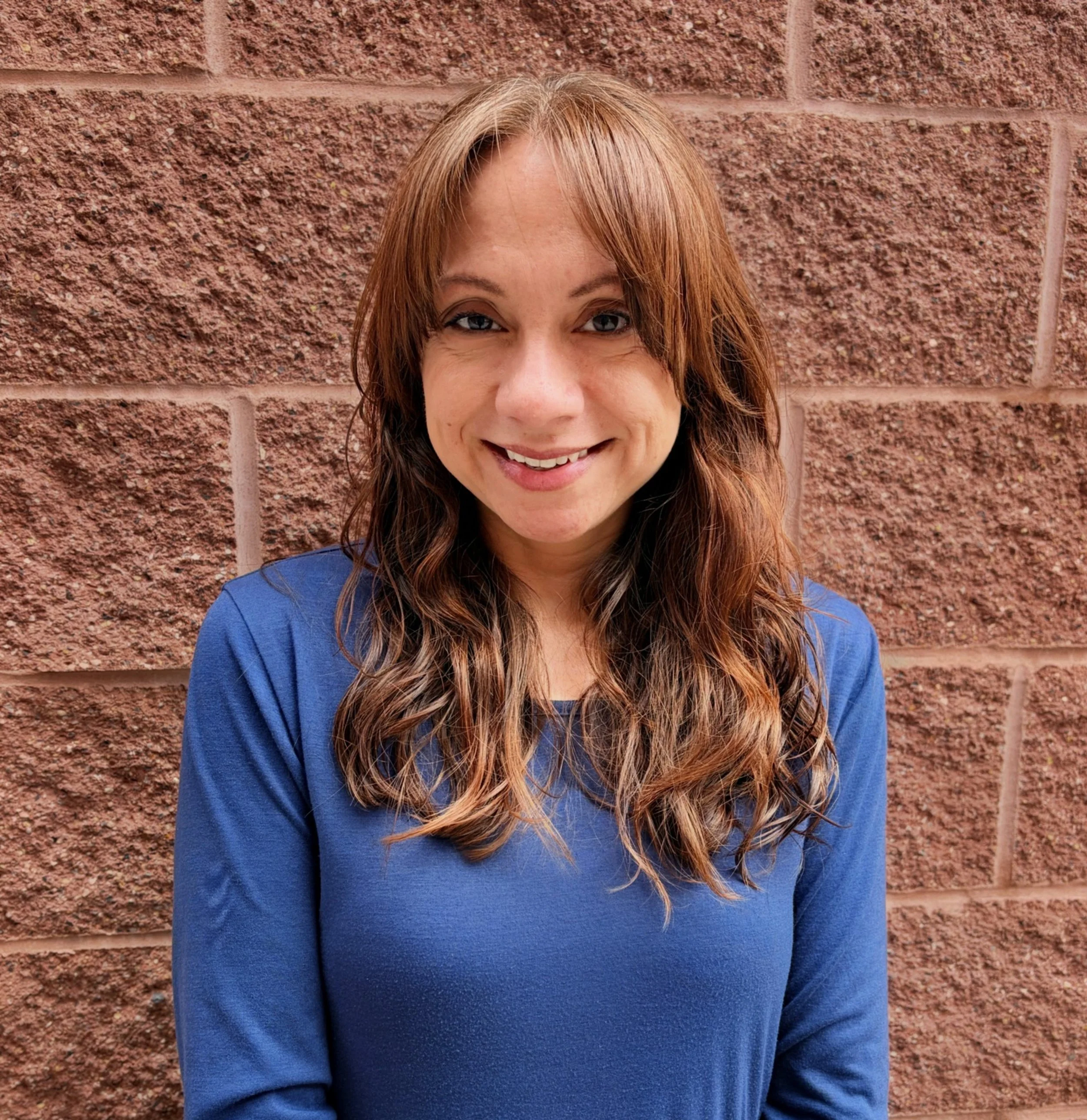 A woman with long, wavy brown hair and light skin, wearing a blue long-sleeve shirt, smiling and standing in front of a red brick wall.