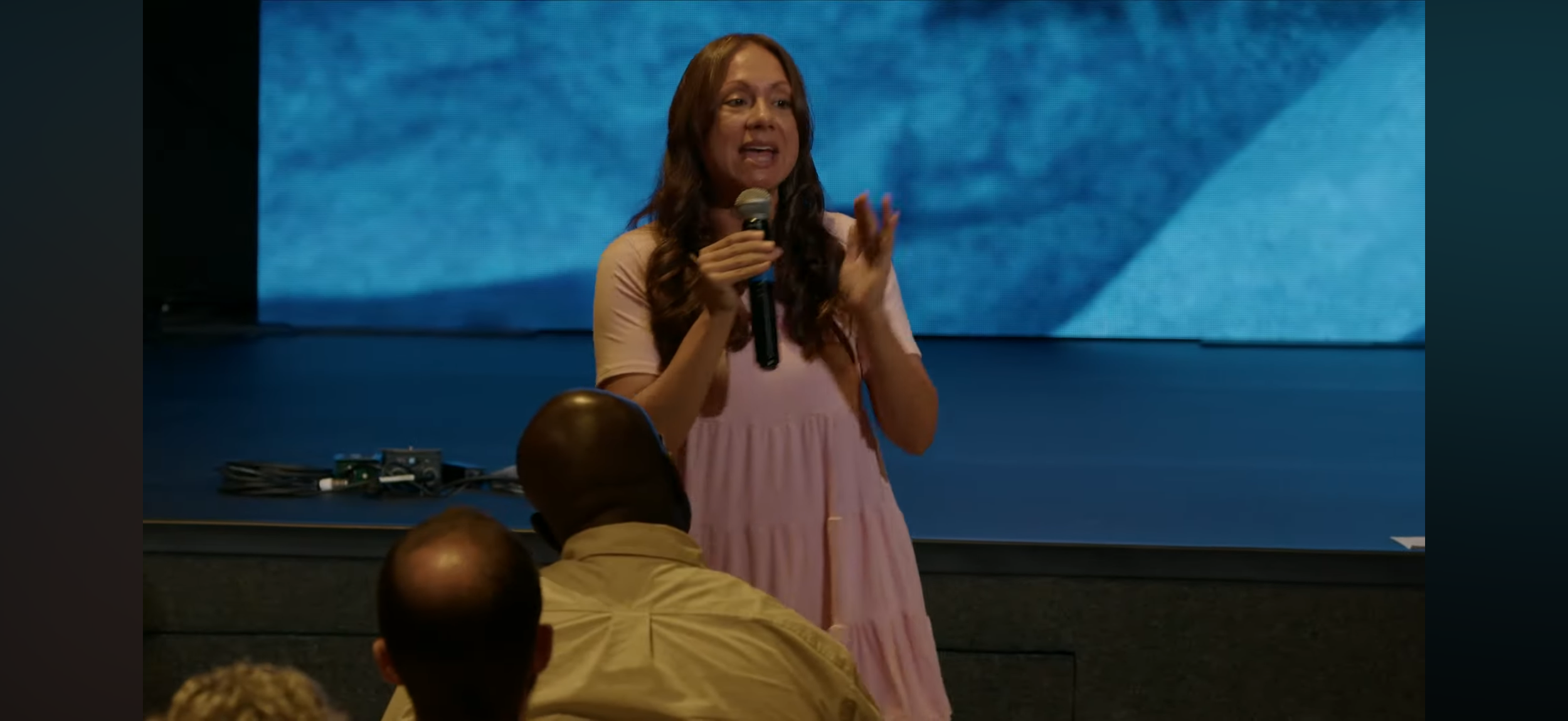 A woman with long brown hair and wearing a pink dress speaking into a microphone in front of an audience, with a blue textured background behind her.