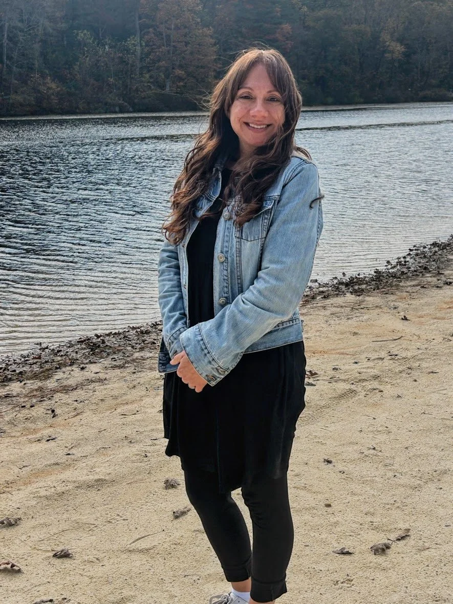 A woman standing on a sandy beach near a body of water with trees in the background, smiling and wearing a denim jacket and black outfit.