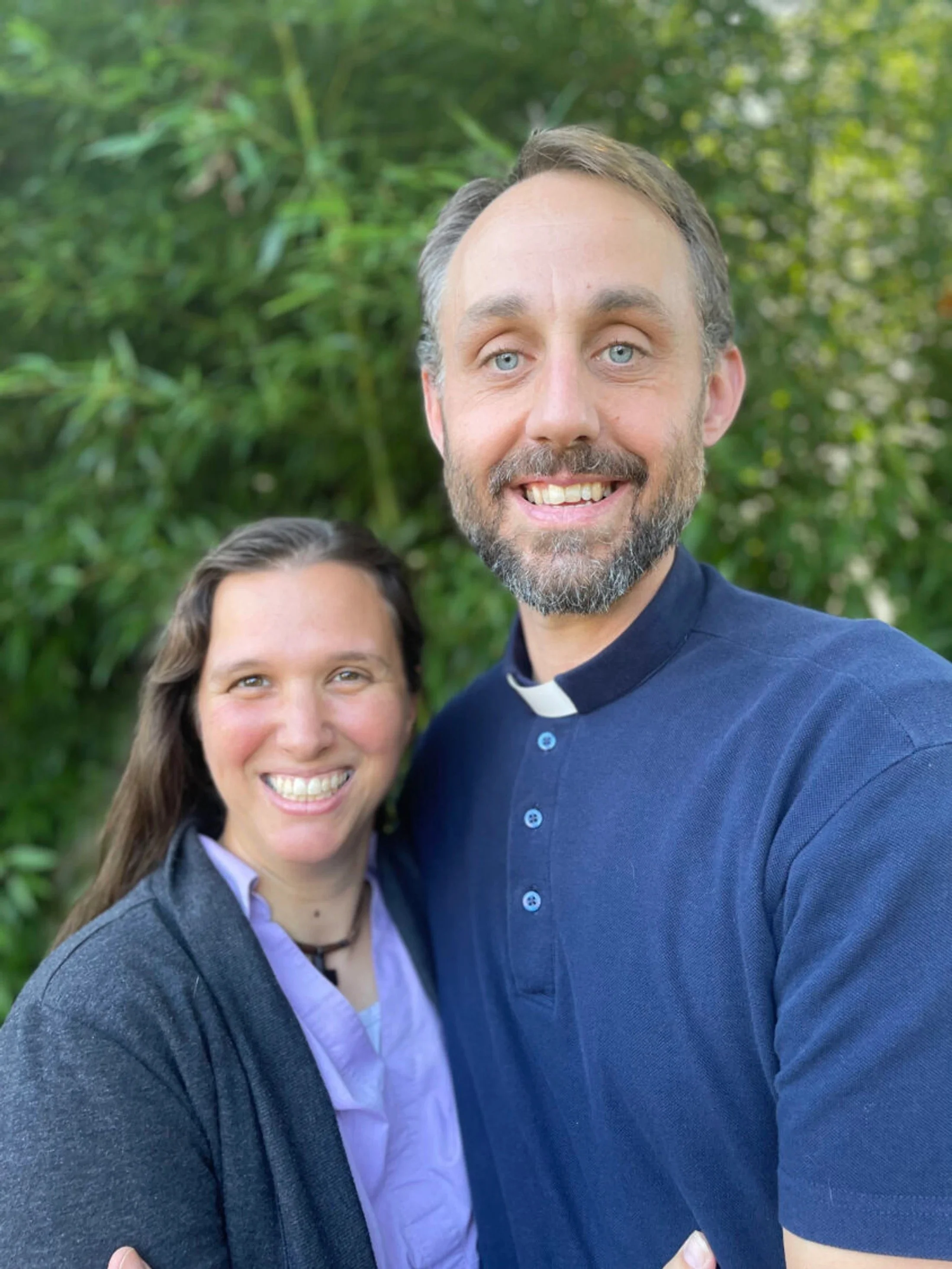 A smiling man and woman taking a selfie outdoors in front of green foliage, both dressed casually.