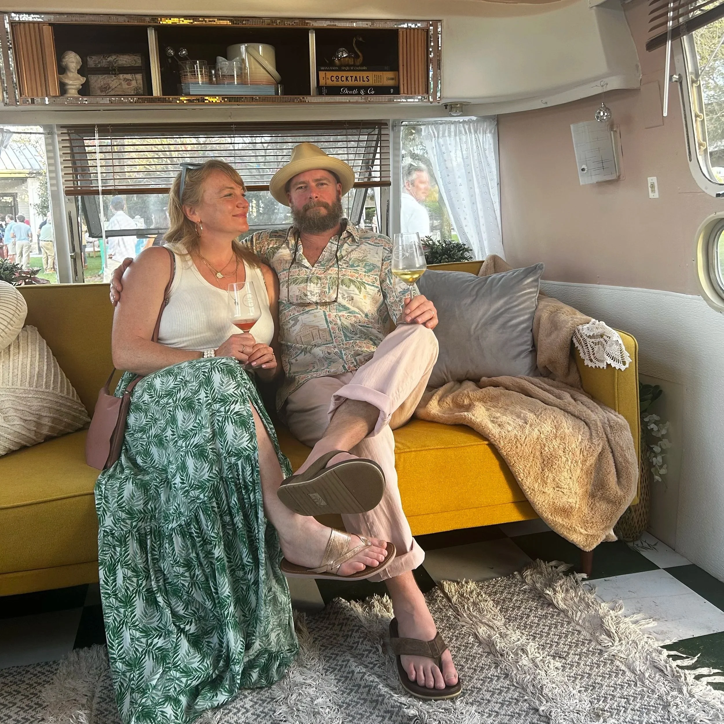 A woman and a man sitting closely together inside a vintage camper trailer. The woman is wearing a white tank top and a patterned green skirt, holding a wine glass, and has earrings. The man has a beard, is wearing a patterned shirt, beige pants, and