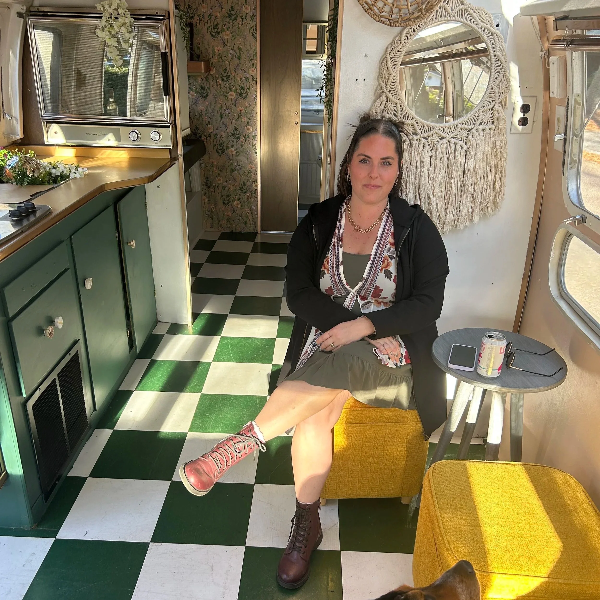 A woman with dark hair and hoop earrings sitting in a vintage camper with retro decor, a large macrame wall hanging, and a window showing an outdoor scene. She is wearing a floral shirt and layered necklaces, with a beverage on a small table in front