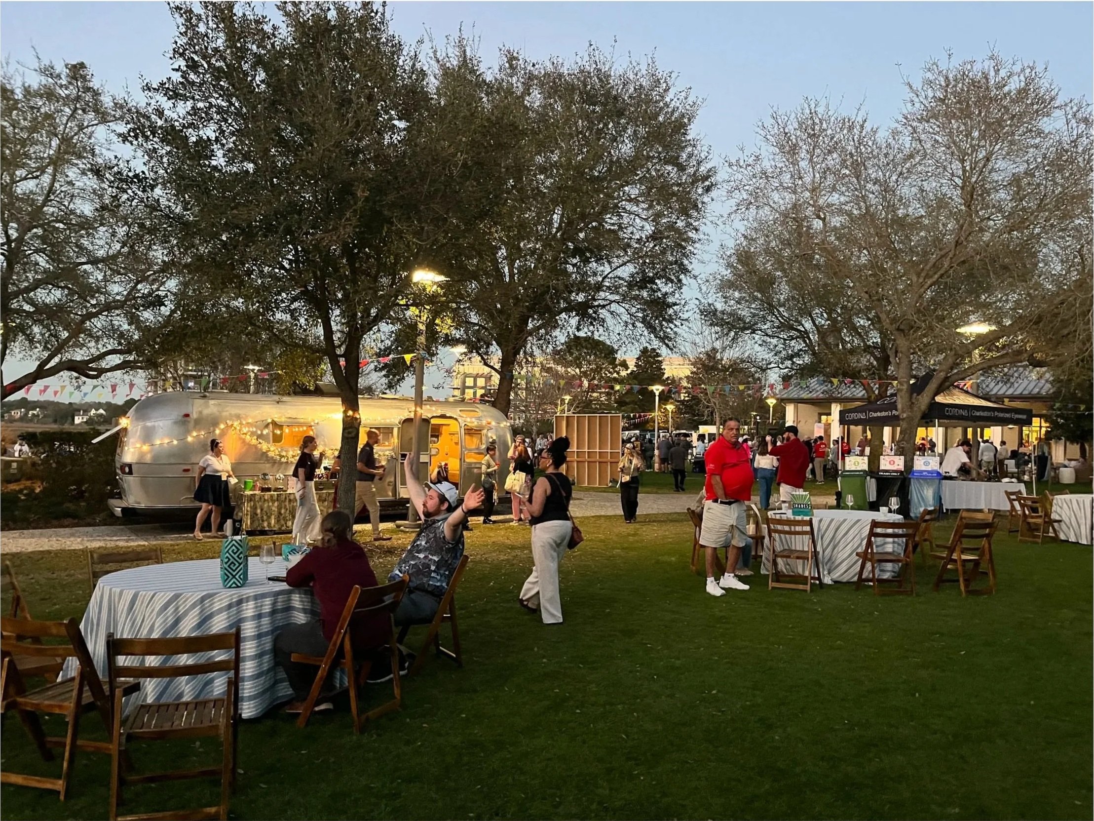 Outdoor evening event with people socializing, trees, string lights, and food trucks in the background.