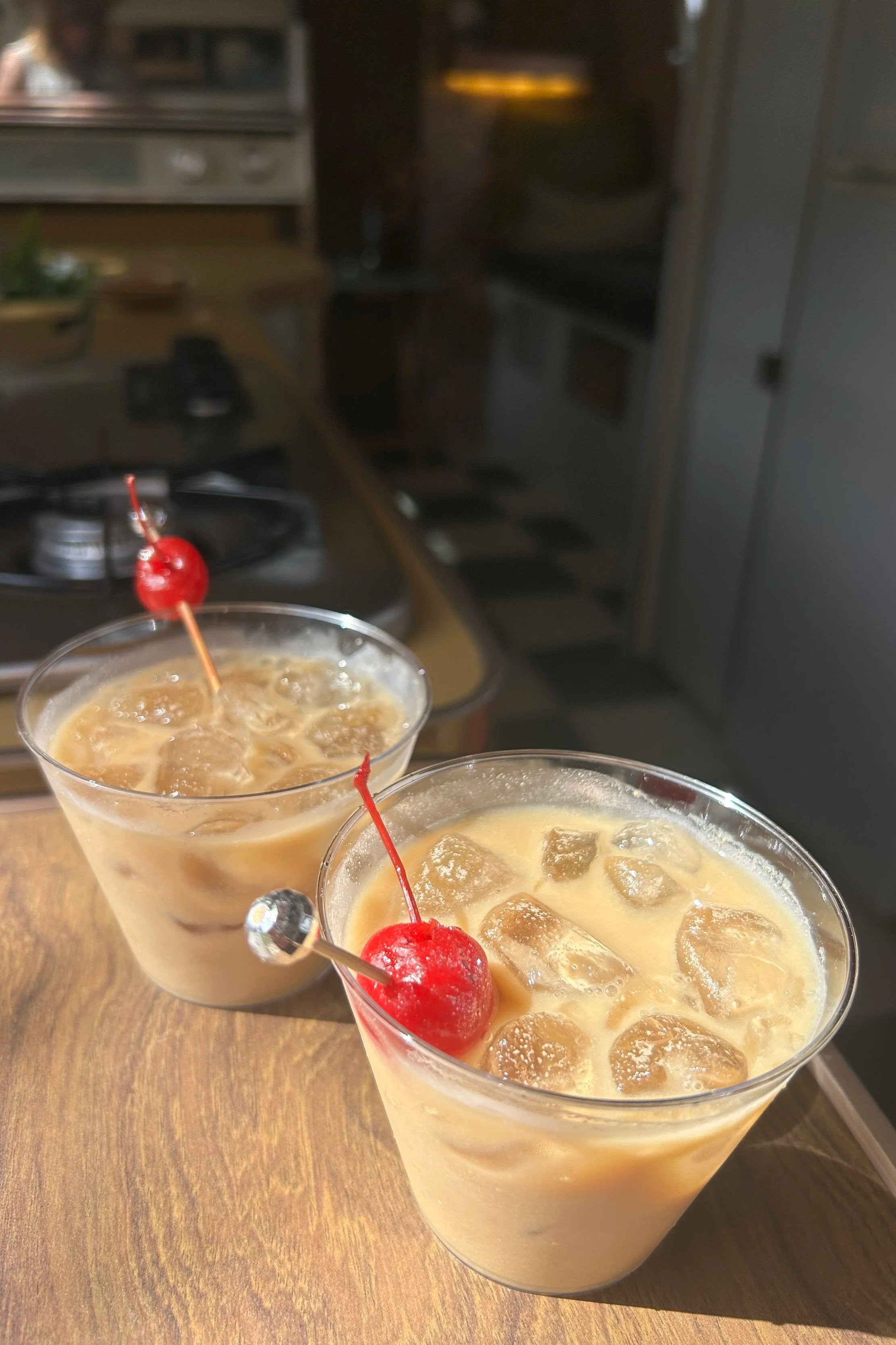Two cocktails with ice and maraschino cherries on a wooden table, in a kitchen setting.