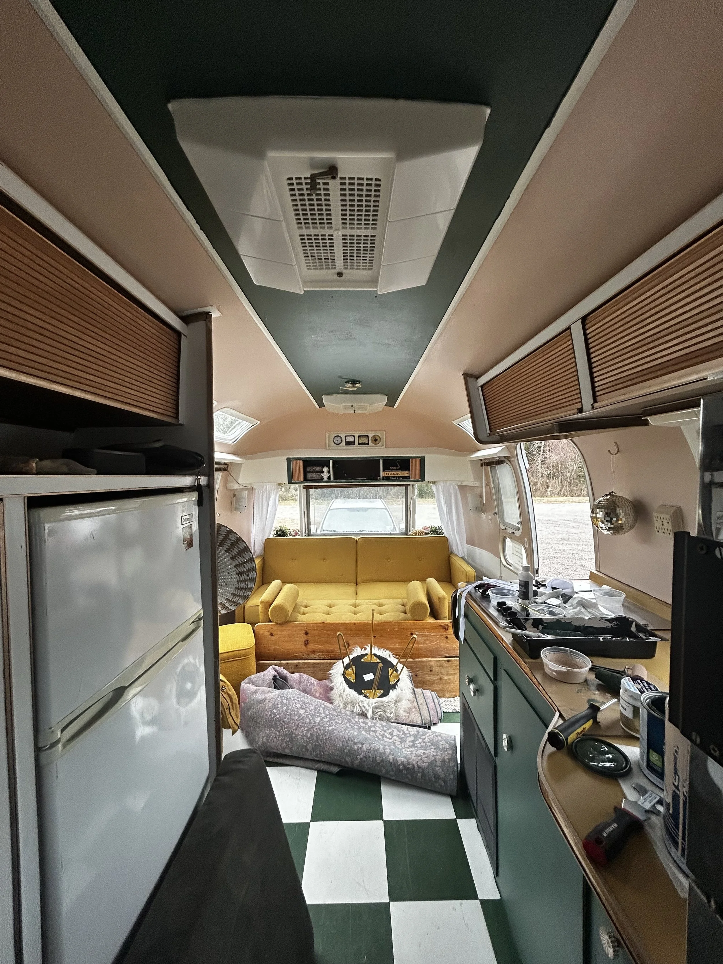 Inside a vintage camper with yellow furniture, a kitchen area on the right, a yellow couch at the back, and a dog laying on a gray blanket in front of the couch. The ceiling has a white vent and a small light fixture. The floor has green and white checkered tiles. There are windows with curtains on either side.