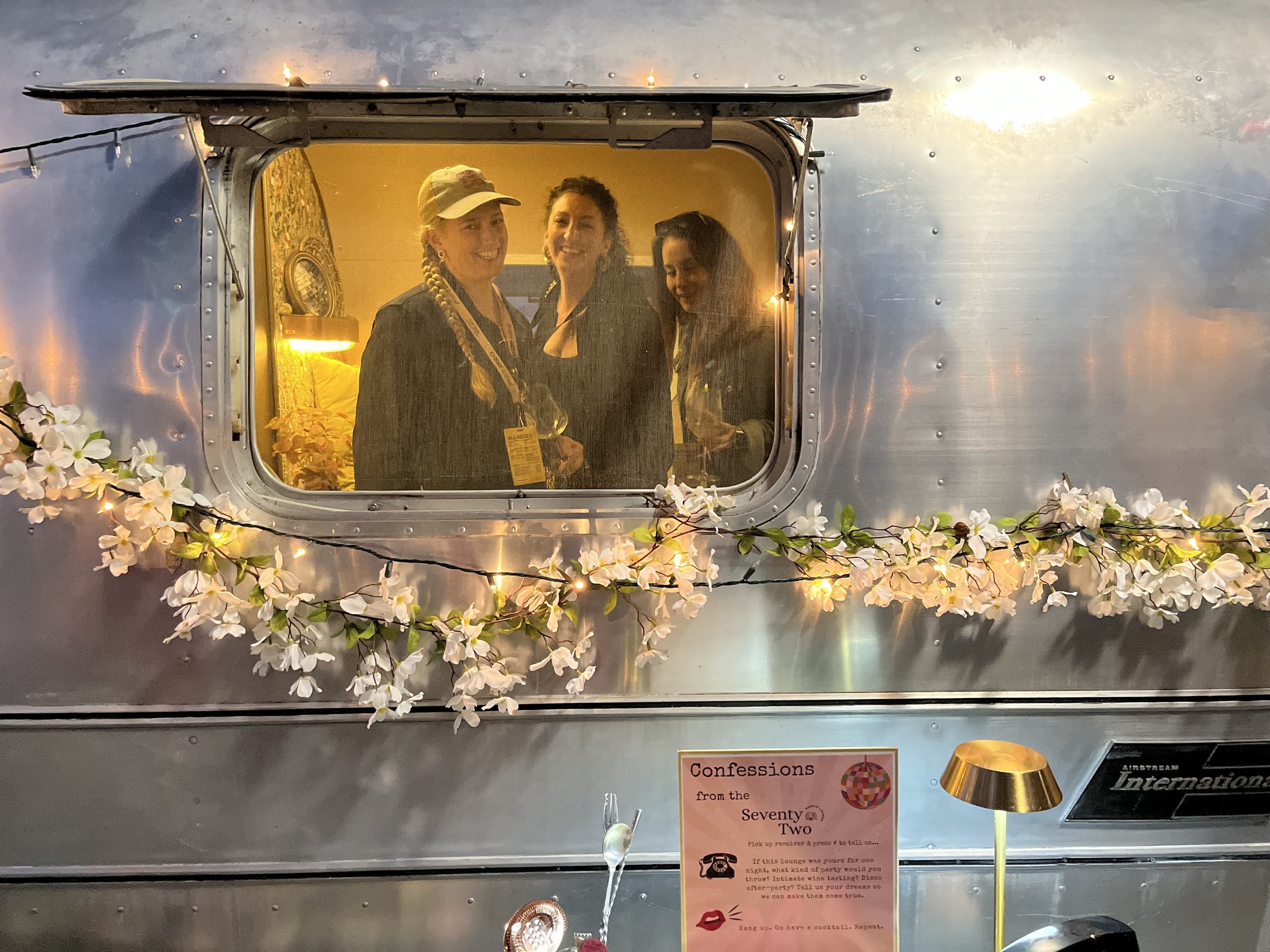 Three women inside a vintage camper trailer, smiling and looking out the window. The trailer is decorated with white flowers and string lights, with a sign below that reads 'Confessions from the Seventy T Two'.