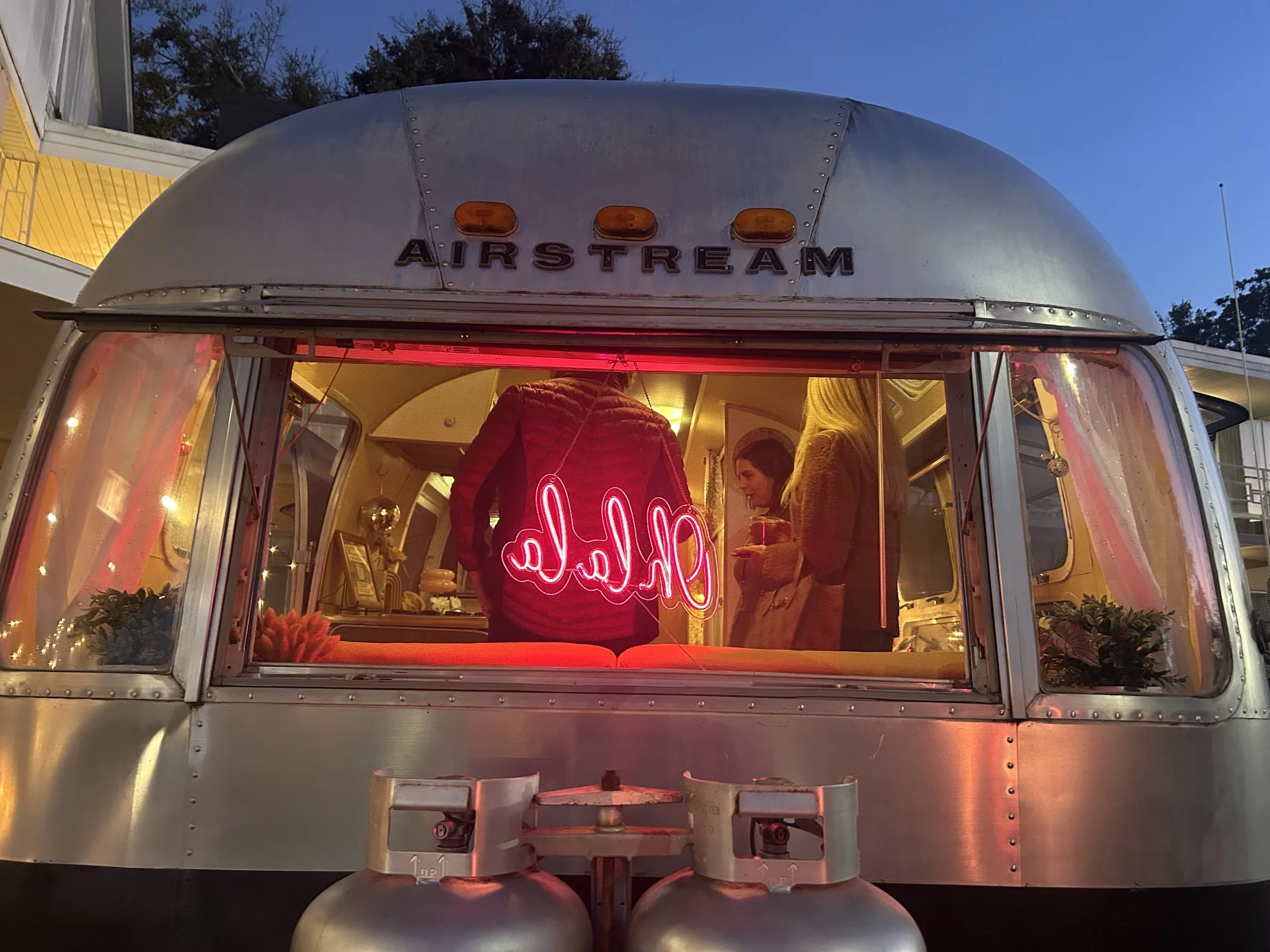 A silver Airstream trailer with the logo on top. The trailer has a large window with pink neon lights that spell out 'hello.' Inside, there are two women, one with blonde hair and another with dark hair, standing and talking. The scene is set during 