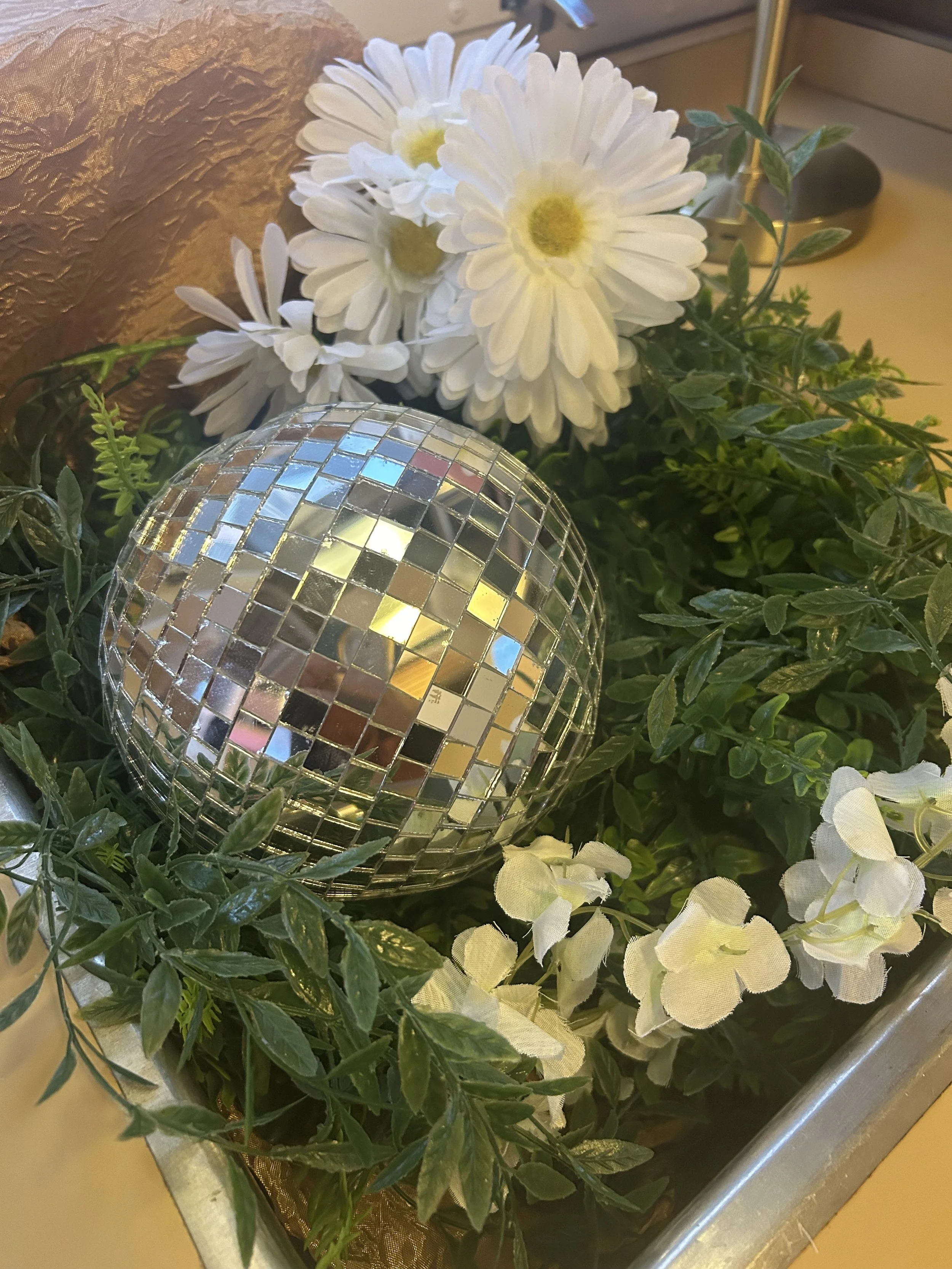 A bouquet of white daisies with yellow centers, surrounded by green foliage, and a decorative disco ball, on a gold fabric background.