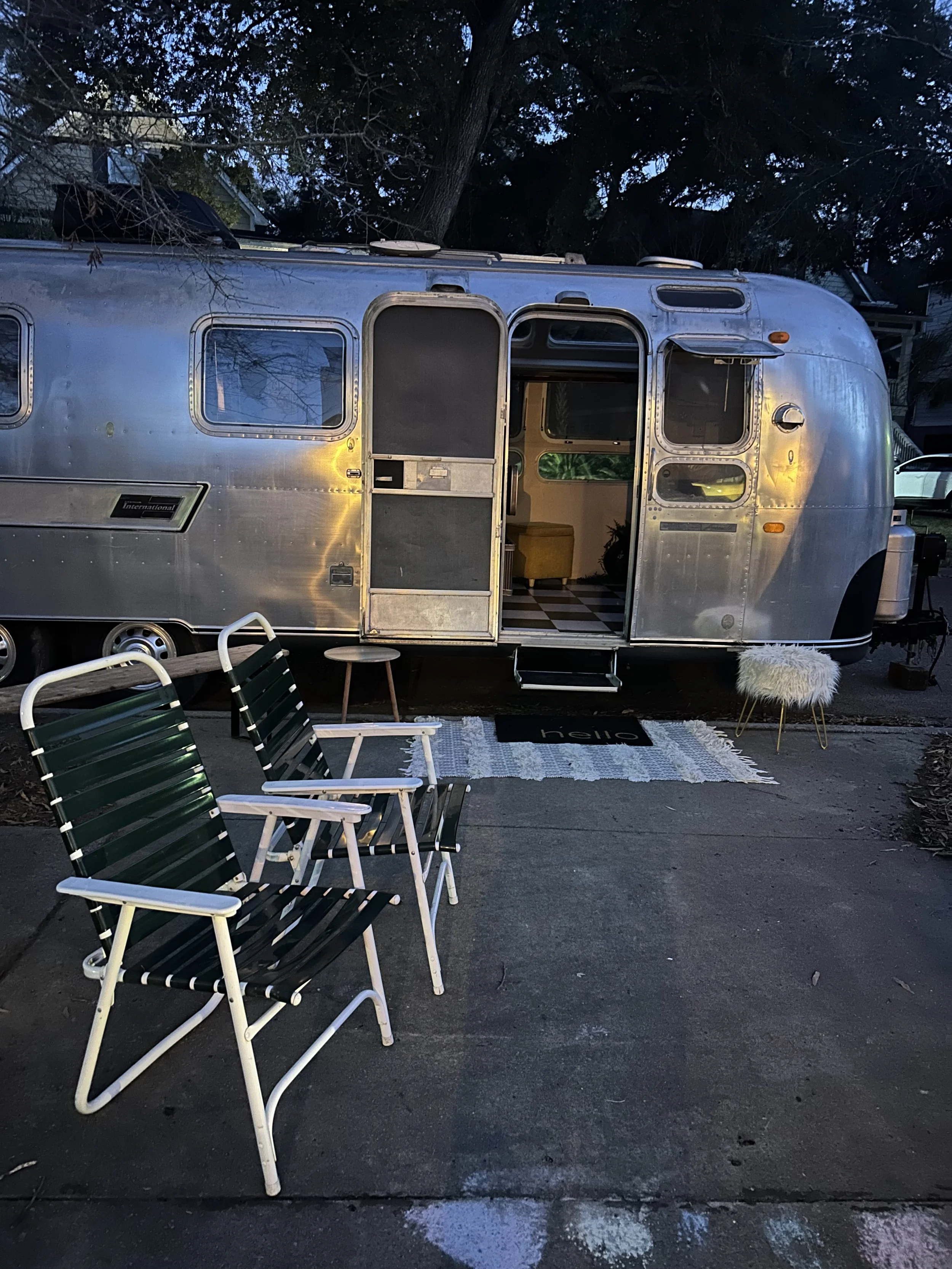 A vintage silver travel trailer parked on a driveway with an open door and a view inside. There are outdoor chairs, a small stool or side table, and a fluffy white stool outside. A welcome mat with the word 'hello' is on the ground in front of the do