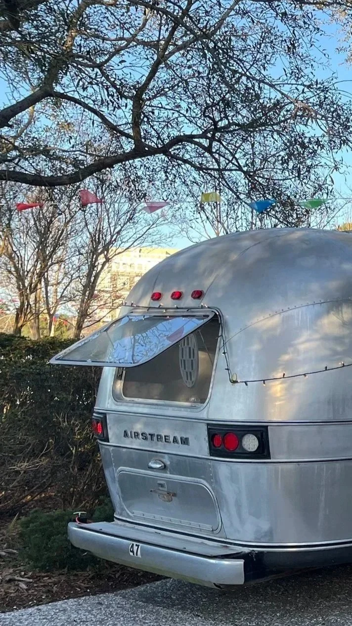Silver Airstream camper trailer parked near bushes with leafless trees and colorful flags hanging overhead in an outdoor setting.