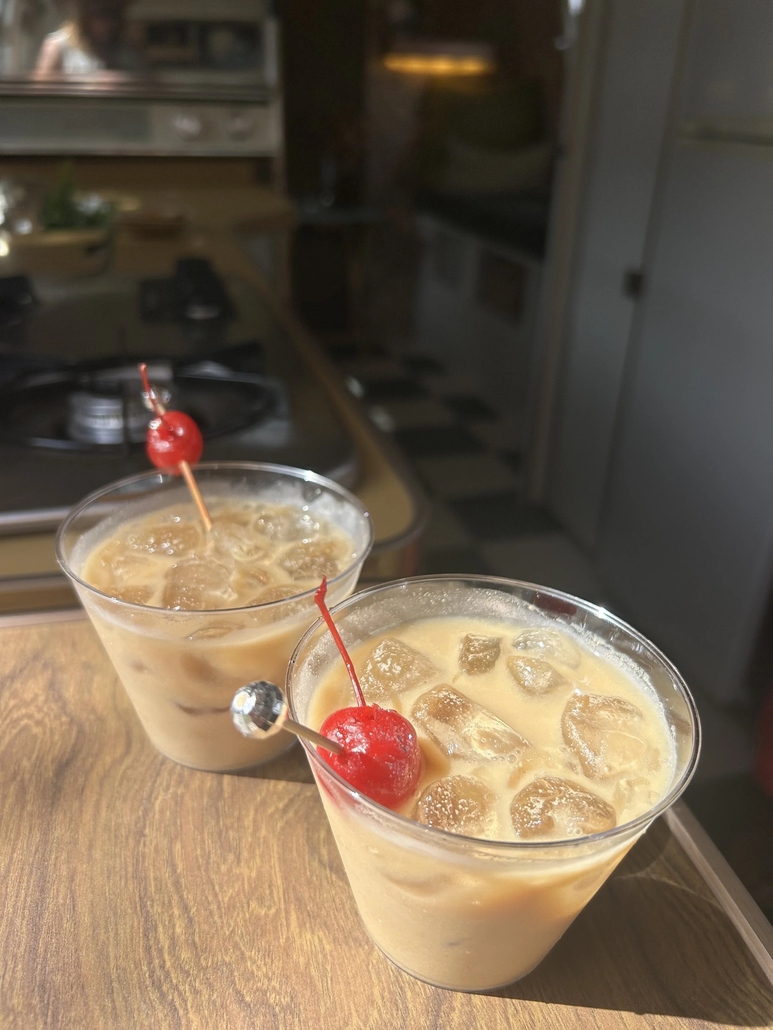 Two cocktails with ice cubes and maraschino cherries on a wooden table, blurred kitchen background.
