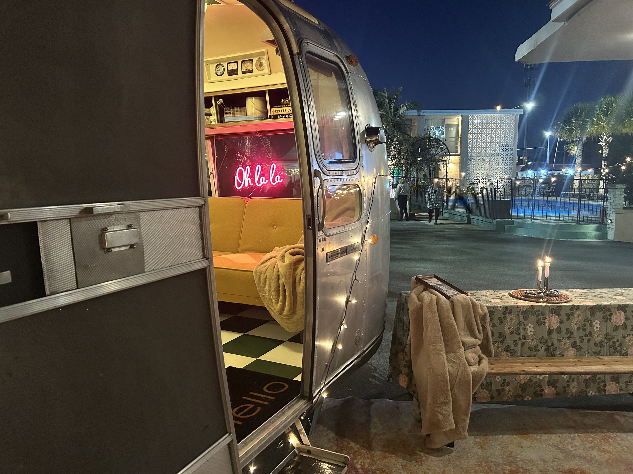 A vintage silver camper trailer with an open door showing a cozy interior with neon 'Oklahoma' sign, yellow seating, and a checkered floor. Outside, there is a table with a lace tablecloth, candles, and blankets. In the background, a pool area with p