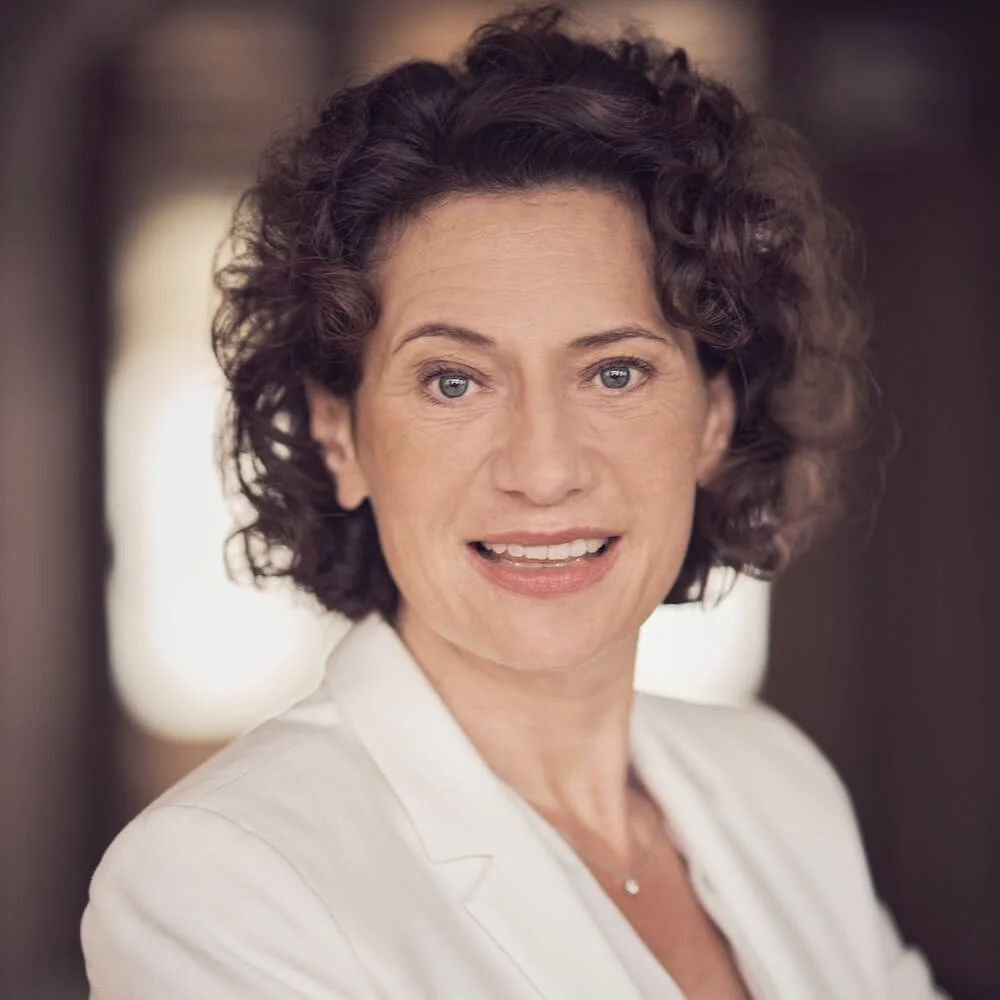 Nahaufnahme von Andrea Belliger, lächelnd, mit dunkelbraunen, lockigen Haaren und weißem Blazer.