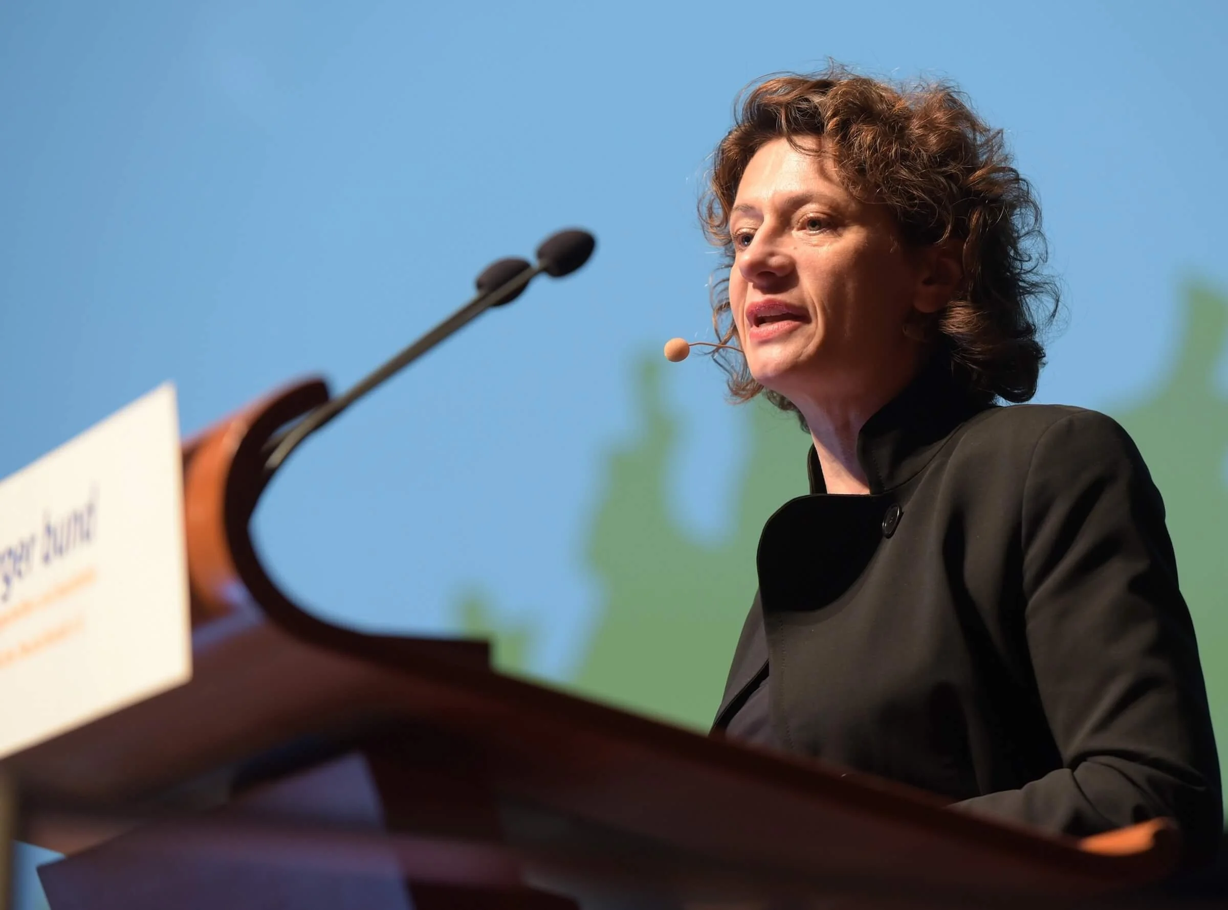 Andrea Belliger spricht an einem Rednerpult bei einer Veranstaltung, mit einem Hintergrund in Blau und Grün.
