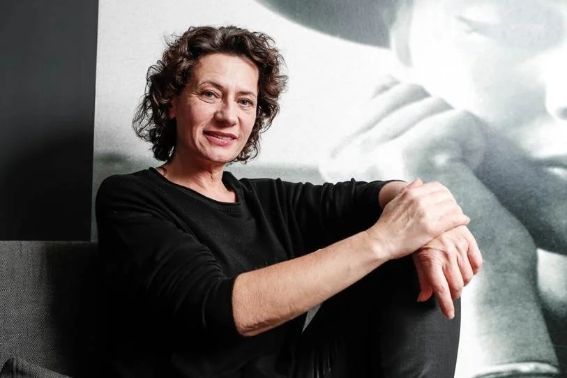 Andrea Belliger sitzend in einem schwarzen Pullover vor einem monochromen Wandbild sitzt, das ein abstraktes Kunstwerk zeigt.