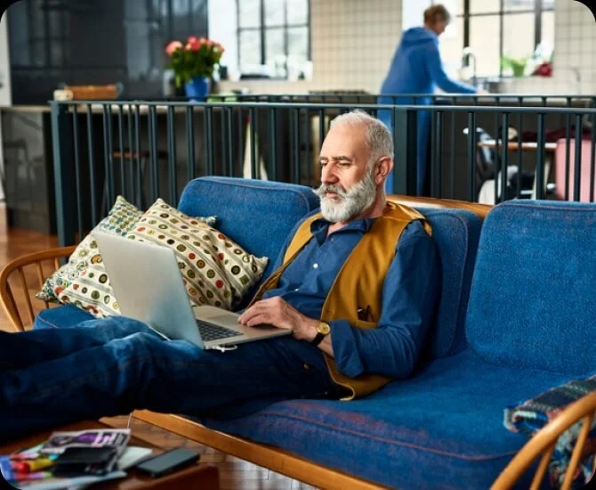Ein älterer Mann mit grauem Bart liegt auf einer blauen Couch, arbeitet an einem Laptop, im Hintergrund eine Frau in blauer Jacke im Büro.