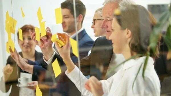 Geschäftsleute arbeiten gemeinsam hinter einer Glasscheibe und kleben gelbe Post-its an die Oberfläche.