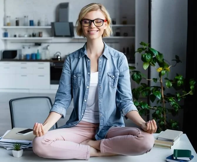 Junge Frau sitzt im Büro, meditiert auf einem Schreibtisch, umgeben von Büromaterialien und Pflanzen, lächelnd.