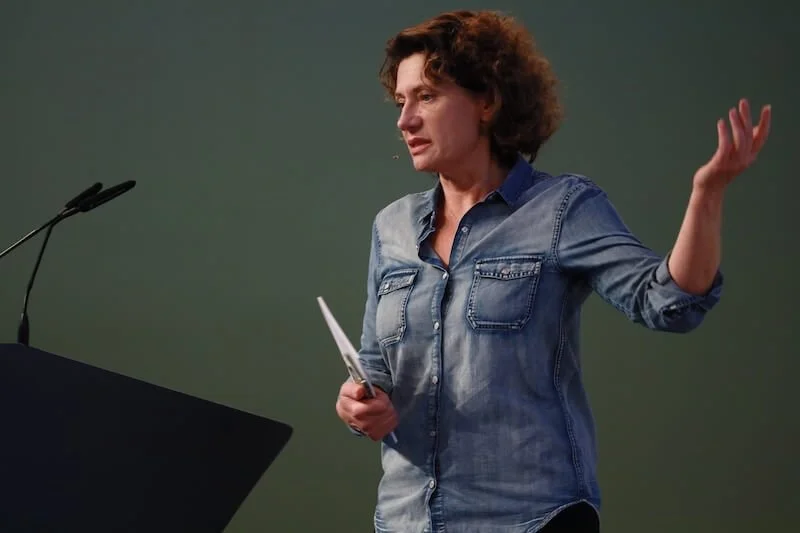 Andrea Belliger an einem Rednerpult, hält einen Zettel in der Hand und gestikuliert während eines Vortrags.