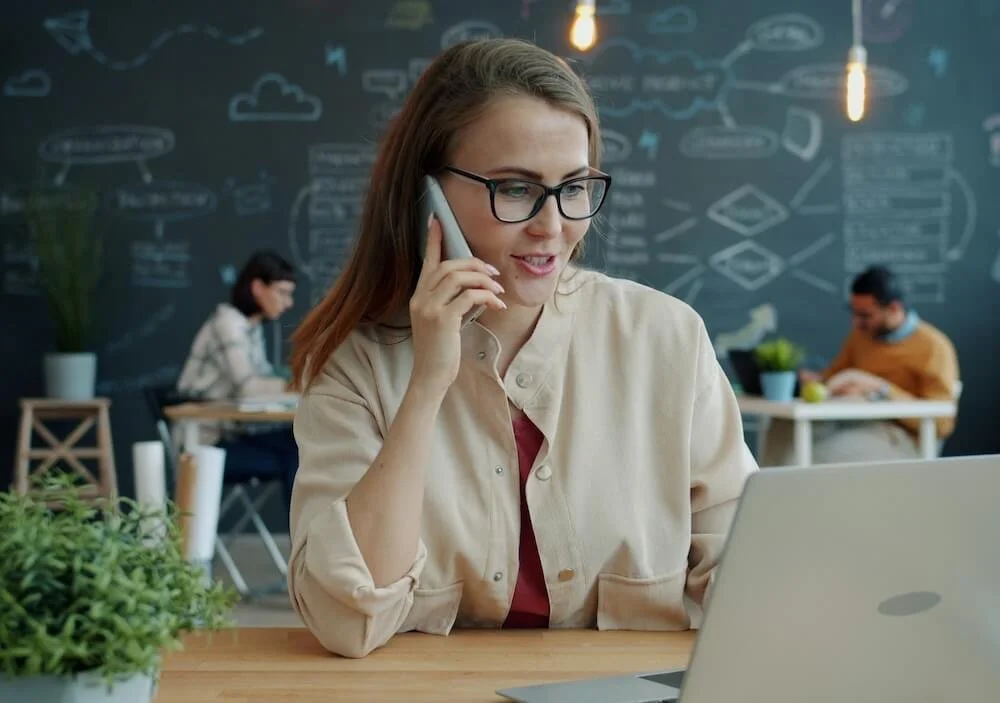 Frau spricht am Telefon, arbeitet am Laptop, im Hintergrund eine Tafel mit Skizzen und Diagrammen, moderne Büroumgebung.