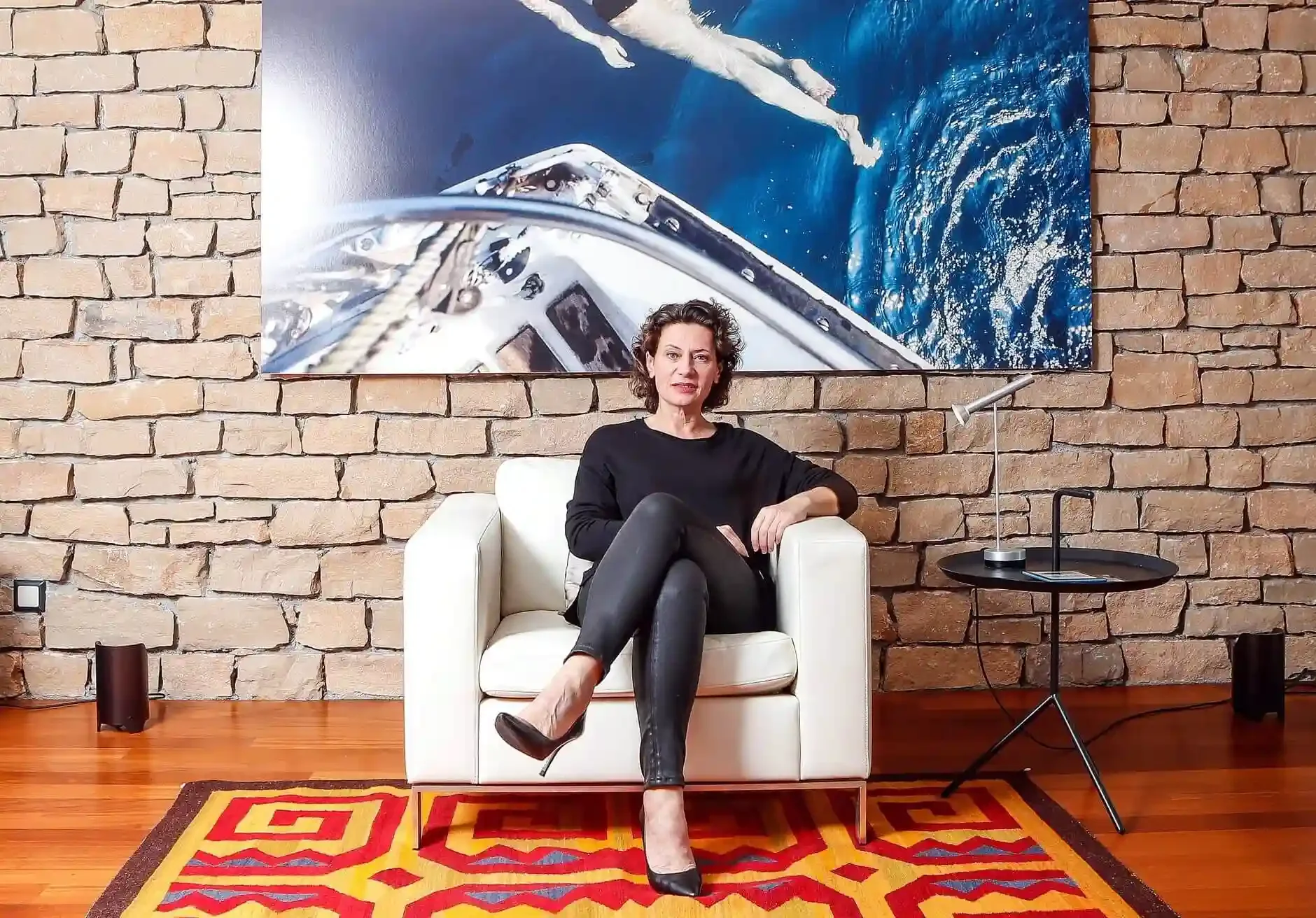 Andrea Belliger sitzt auf einem weißen Sessel vor einer Backsteinwand mit einem großen Wandbild eines Bootes auf dem Wasser.