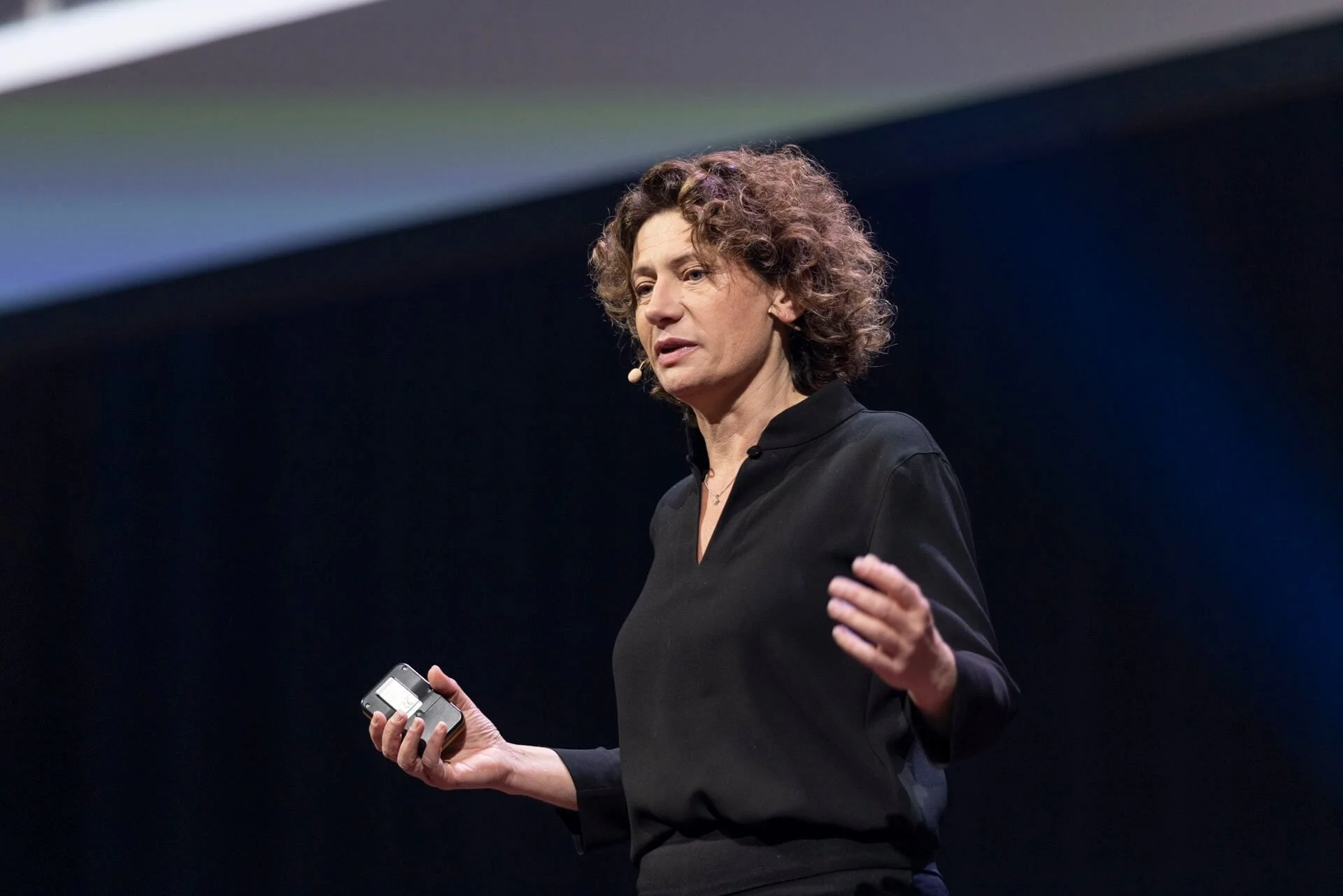 Andrea Belliger mit lockigem Haar hält ein Mikrofon in der rechten Hand und spricht auf einer Bühne.