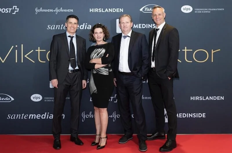 Andrea Belliger mit drei Männern, alle in formeller Kleidung, posieren vor einem dunklen Hintergrund mit Unternehmens- und Sponsoring-Logos auf einer roten Teppichfläche.