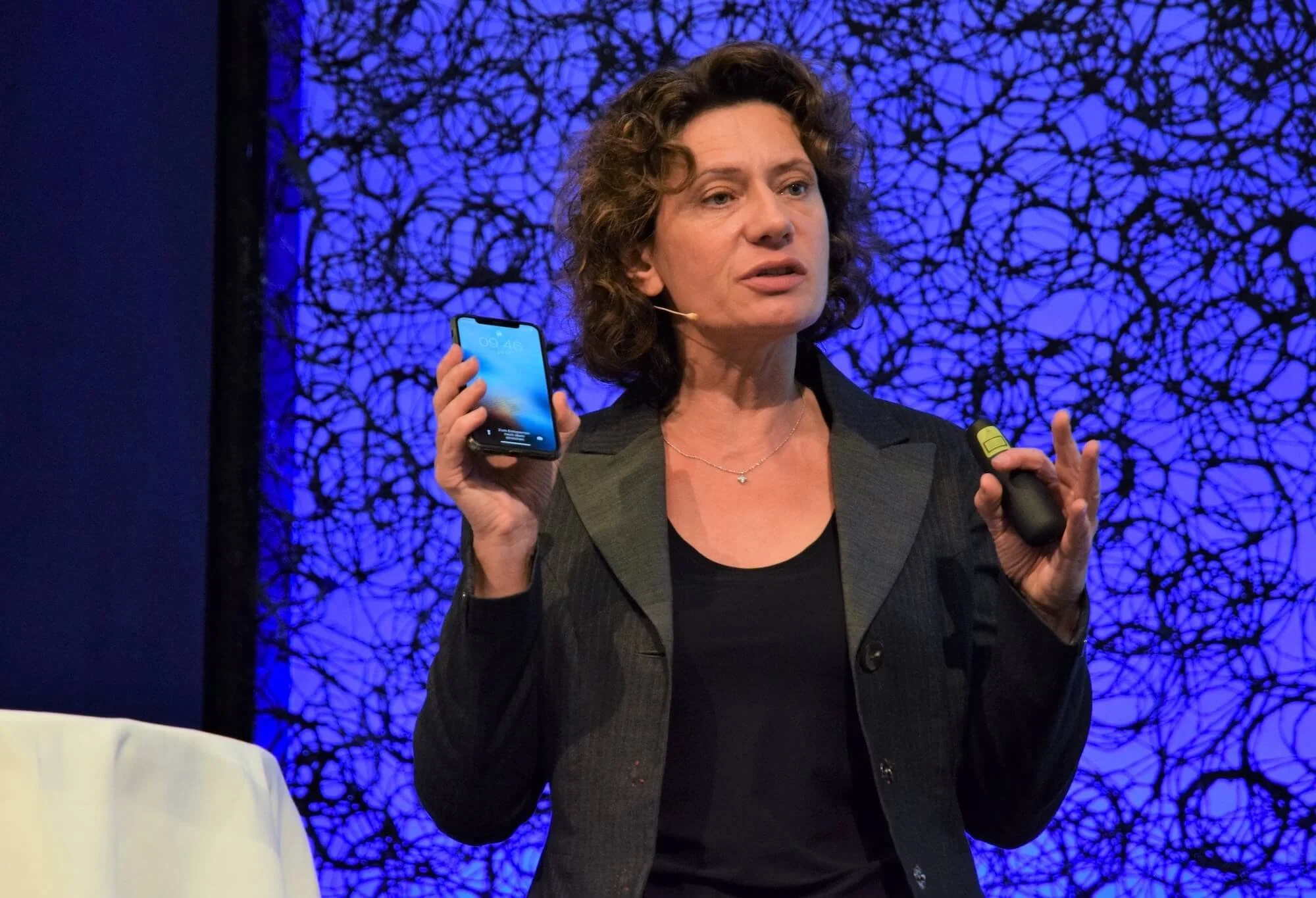 Andrea Belliger bei einer Präsentation mit einem Smartphone in der rechten Hand, vor einem blauen, abstrakten Hintergrund.