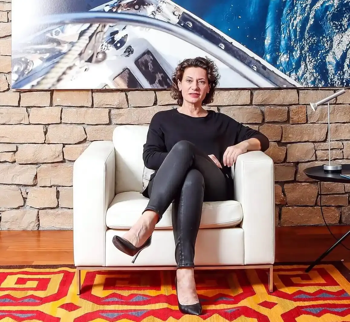 Andrea Belliger sitzt auf einem weißen Sessel vor einer Ziegelmauer, im Hintergrund ist ein Wandbild eines Bootes auf dem Wasser zu sehen.