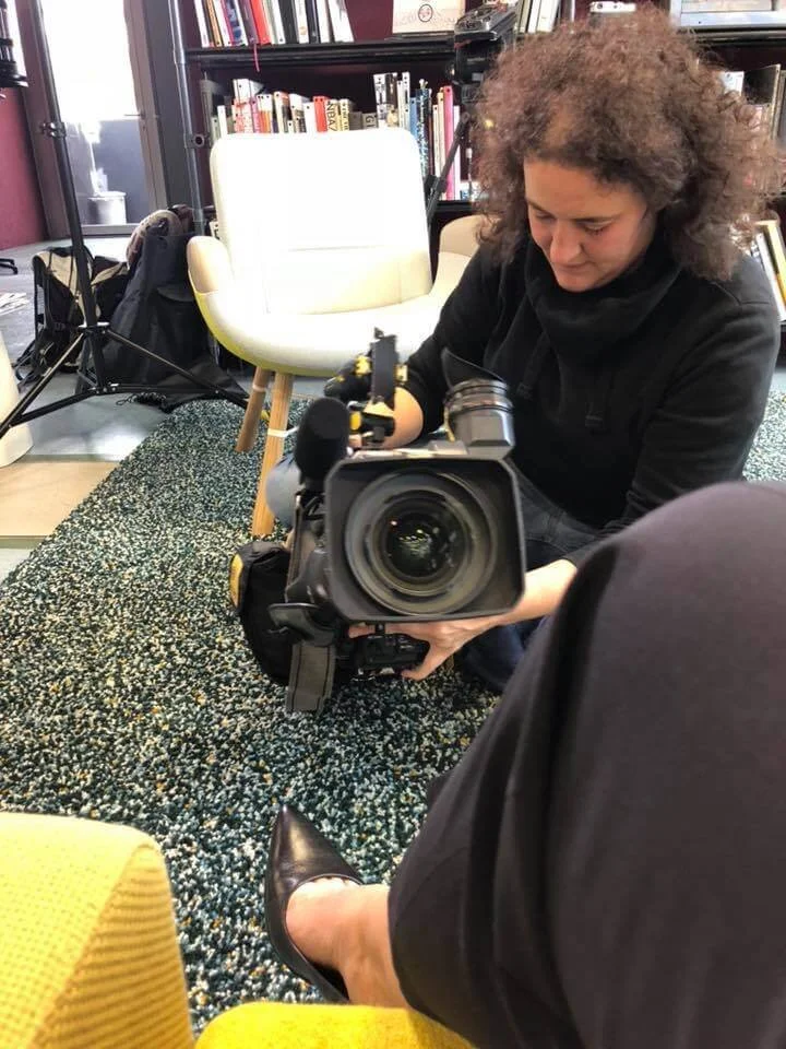 Eine Frau mit lockigem Haar, die eine Kamera hält und sich auf dem Boden befindet, im Hintergrund Bücherregale und ein weißer Sessel in einem Büro.