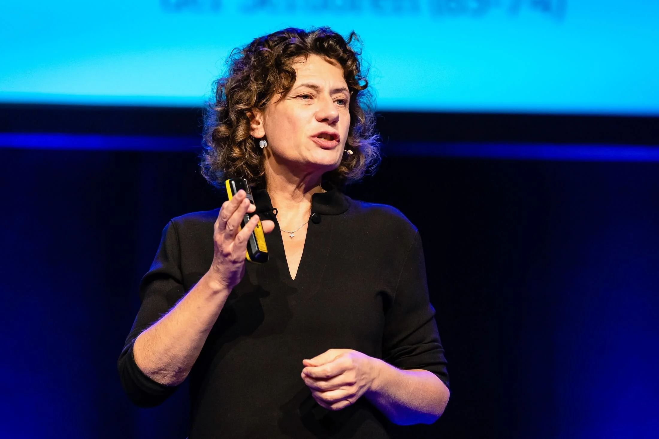 Andrea Belliger hält ein Mikrofon in der rechten Hand und spricht bei einer Veranstaltung vor einem blauen Hintergrund.