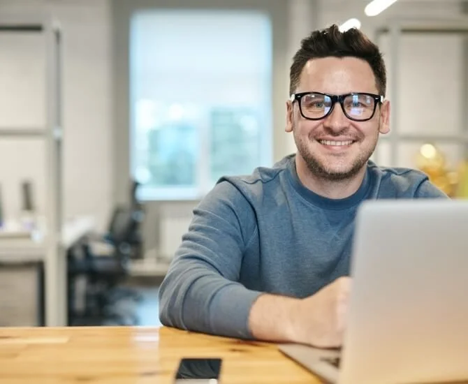 Ein Mann mit Brille, lächelnd, sitzt an einem Tisch im Büro mit einem Laptop.