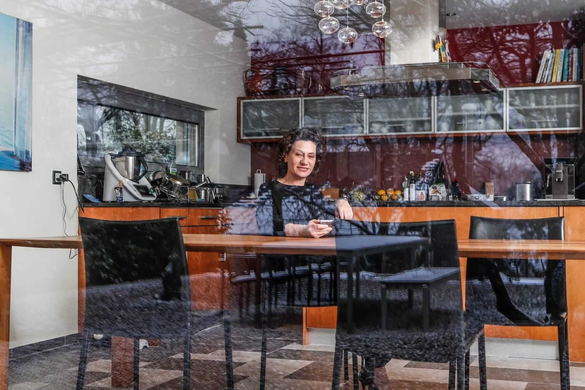 Andrea Belliger sitzt an einem Esstisch in einer modernen Küche, fotografiert durch eine Fensterfront mit Reflexion von Bäumen im Glas.