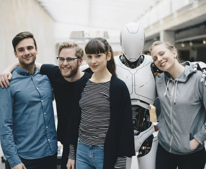 Vier Menschen und ein humanoider Roboter stehen zusammen und lächeln in einem modernen Gebäude.