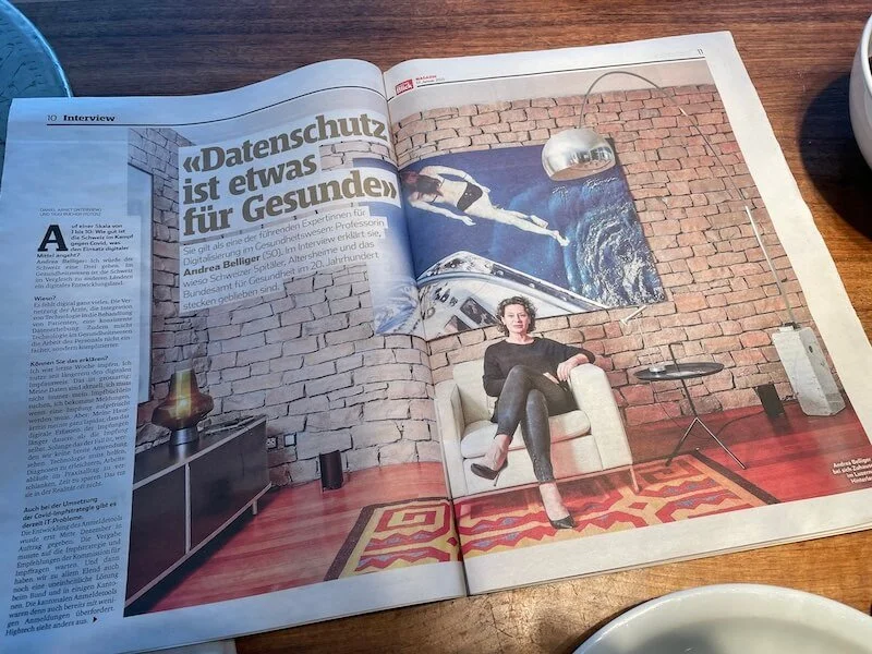 Offene Zeitung mit einem Interview-Artikel, auf dem eine moderne, stilvolle Wohnzimmerwand mit einem Gemälde eines Astronauten im Weltraum und einem proteinschräg an der Wand hängt, sowie Andrea Belliger , die auf einem weißen Sessel sitzt.