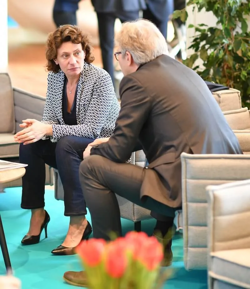 Andrea Belliger und ein Mann sitzen in einem Gespräch in einem modernen Raum, im Hintergrund Pflanzen.