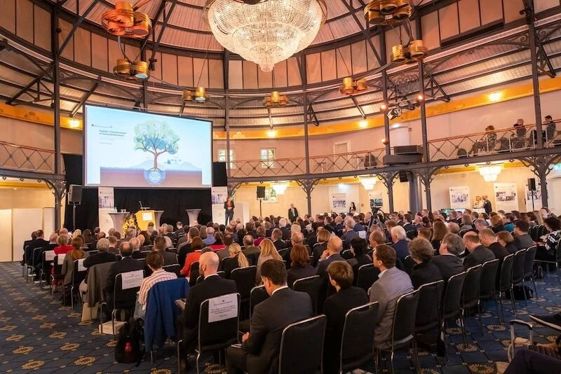 Ein großer Saal mit vielen Menschen, die an einem Konferenz- oder Podiumsgespräch teilnehmen, mit einem großen Bildschirm und einer Deckenleuchte.