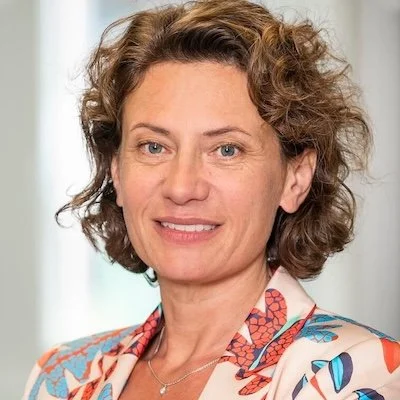 Porträt von Andrea Belliger, die einen bunten Blazer trägt, vor einem hellen Hintergrund.