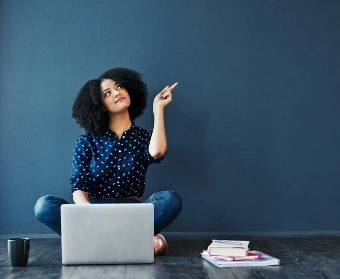 Eine junge Frau mit lockigem schwarzen Haar sitzt auf dem Boden vor einem Laptop, denkt nach und zeigt nach oben.