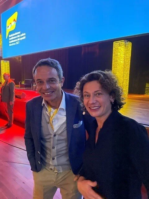 Andrea Belliger und ein Mann posieren auf einer Bühne bei einer Veranstaltung. Im Hintergrund ein Bildschirm mit einem Logo und Text auf Deutsch. Die Umgebung ist bunt beleuchtet.