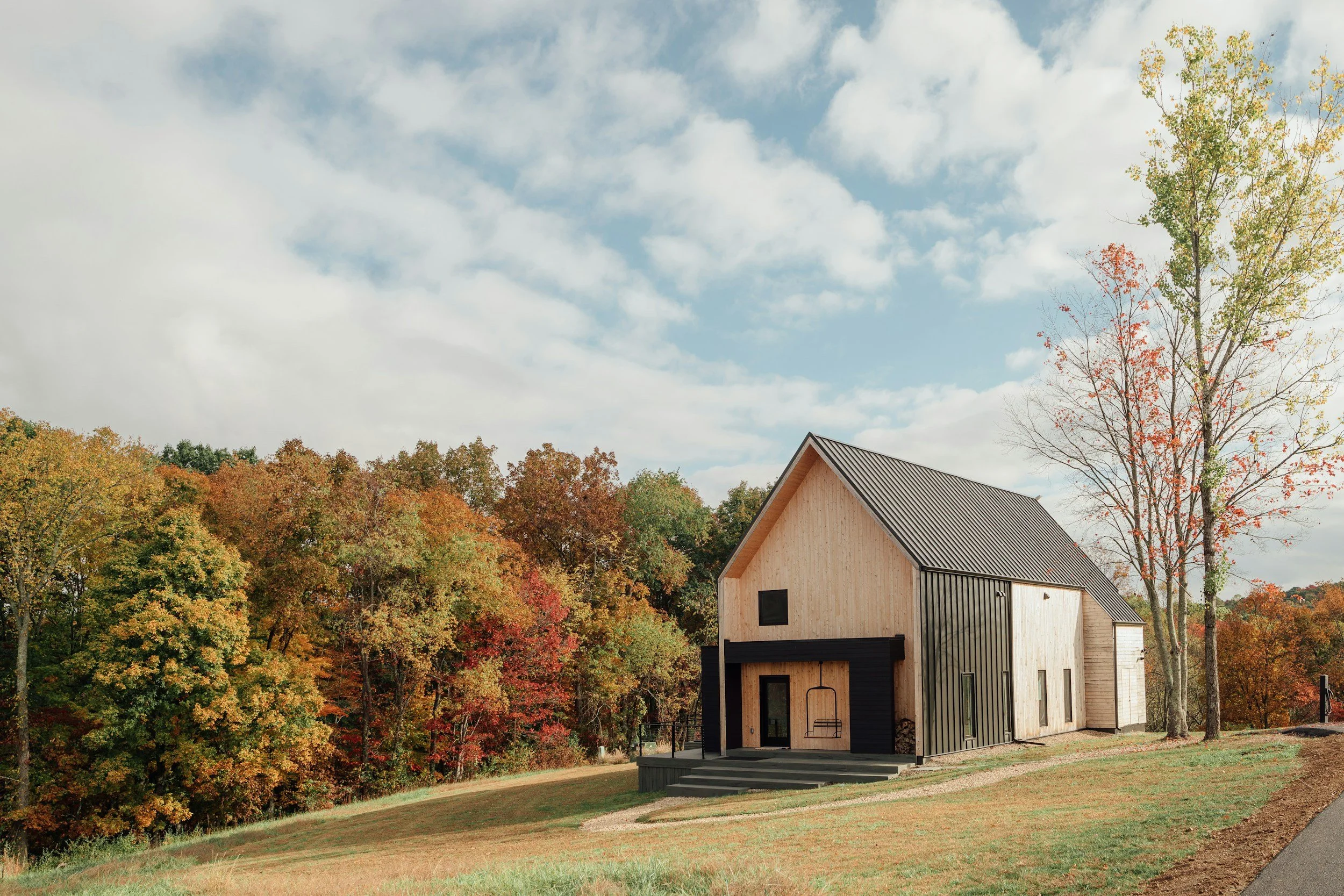 A modern house with a black and natural wood exterior, set on a grassy slope with fall-colored trees in the background under a partly cloudy sky.